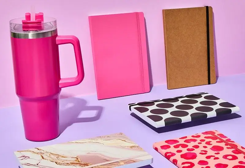 Collection of colorful stationery items and a bright pink travel mug, including notebooks with pink, kraft, polka dot, marble, and leopard print covers